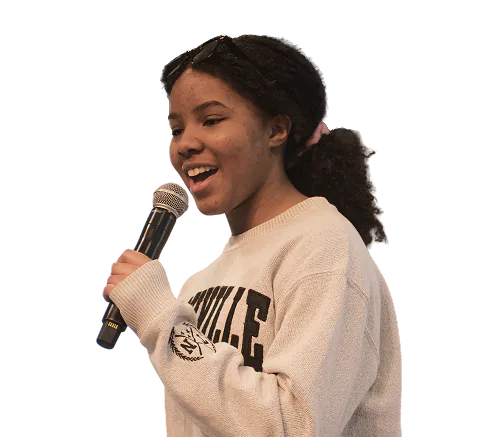 Young woman with curly hair tied back and glasses on her head speaking into a microphone, wearing a light-colored sweater.